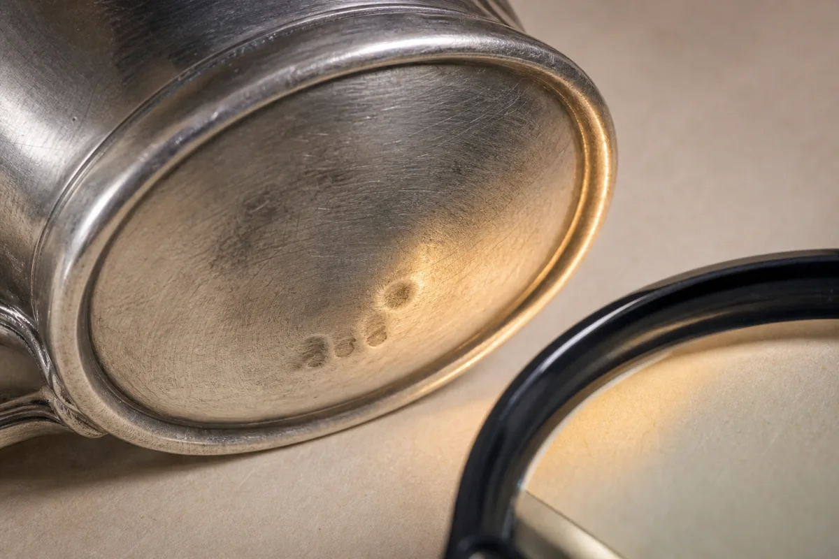 Inspecting marks on a silver pitcher Close illustrated view of faint, unreadable marks near the foot of a silver pitcher, seen beside a magnifier.